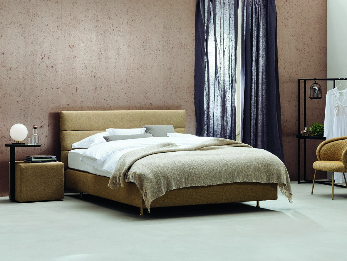 Schramm Boxspring Bett in Beige im Hintergrund Wand mit Holzstruktur und Vorhang